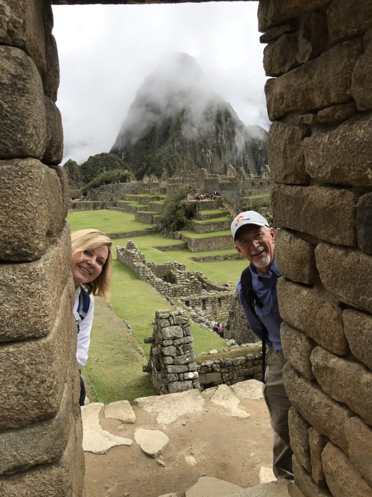 Two tourists smiling and peeking through an ancient stone doorway at Machu Picchu.