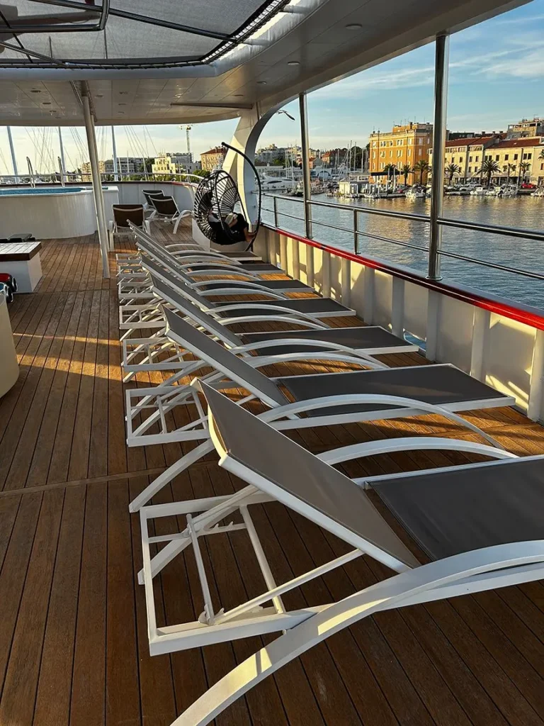 Empty lounge chairs on a ship deck overlooking the water at sunset.